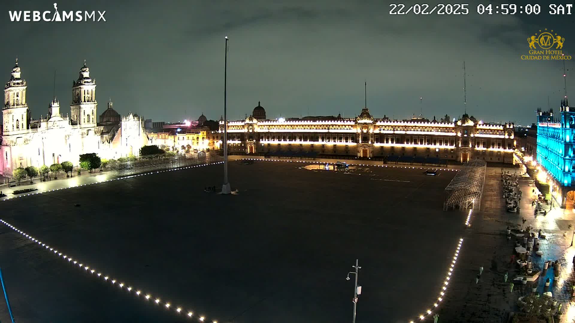 A large, mostly empty plaza at night, illuminated by lights, with a brightly lit cathedral and palace visible in the background under an overcast sky.