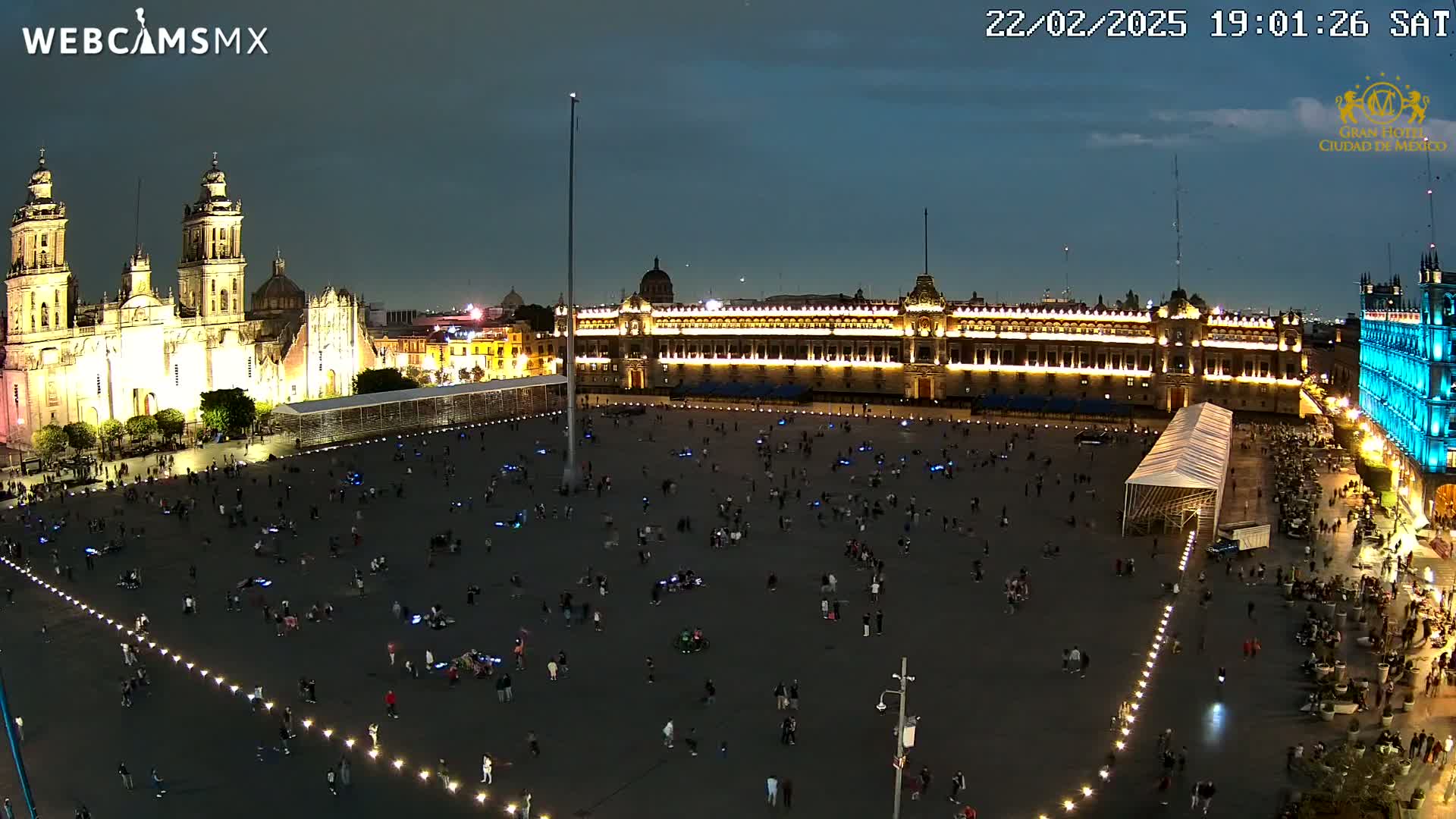 A large, mostly empty plaza at night is illuminated, surrounded by brightly lit buildings, including a cathedral and palace, under a partly cloudy sky.