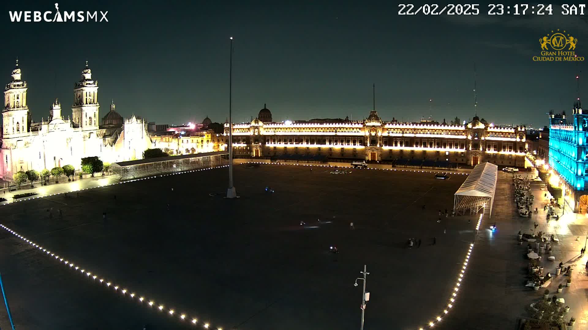 A large, dark plaza at night is illuminated by lights outlining its edges, with a brightly lit cathedral and palace visible in the background.