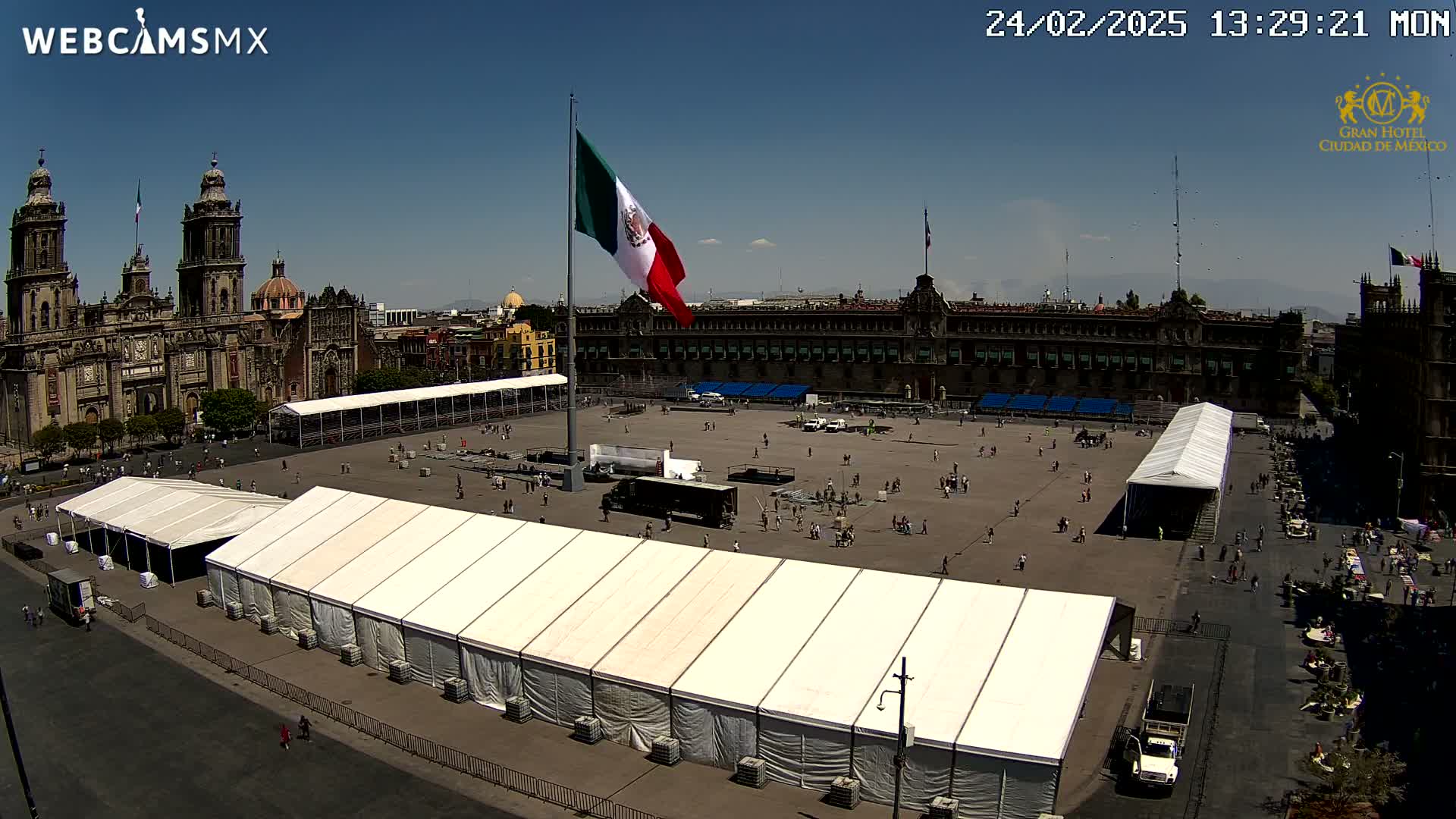 A large plaza, under a clear sunny sky, contains several white tents and a large Mexican flag on a tall pole, with a cathedral and government buildings in the background.
