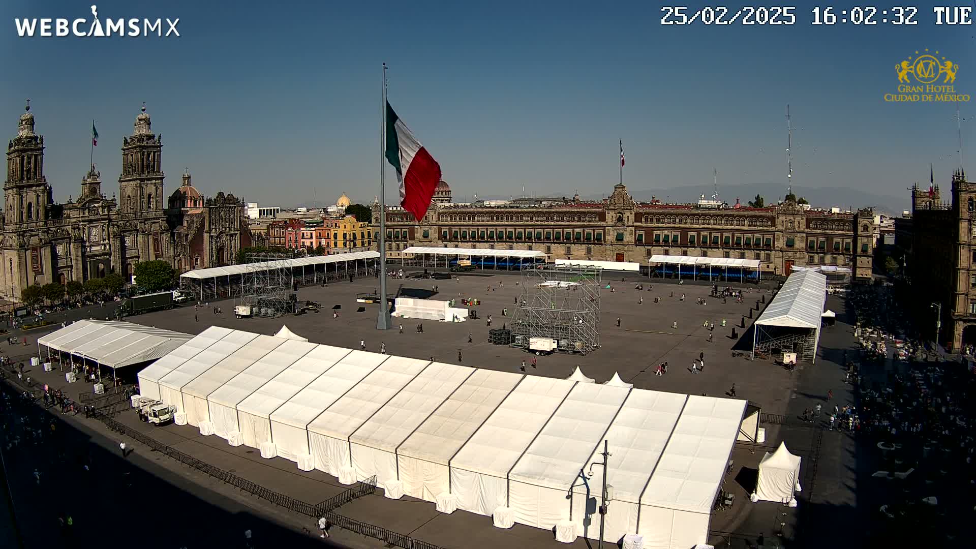 A large plaza, under sunny skies, features several white tents, scaffolding, and a Mexican flag flying high near a cathedral and government building.