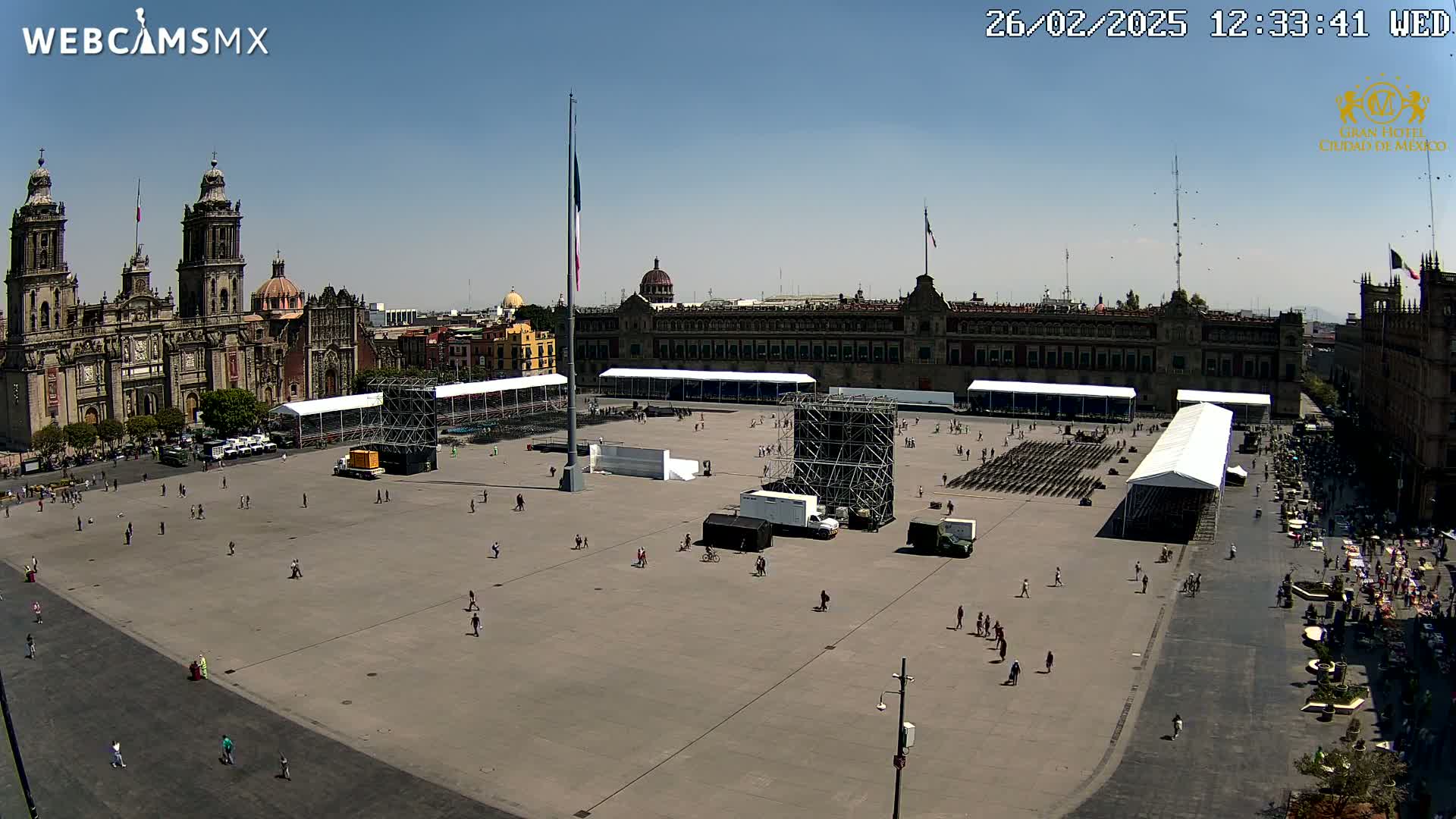 A large plaza with a few people, temporary structures, and a large building in the background under a clear blue sky.
