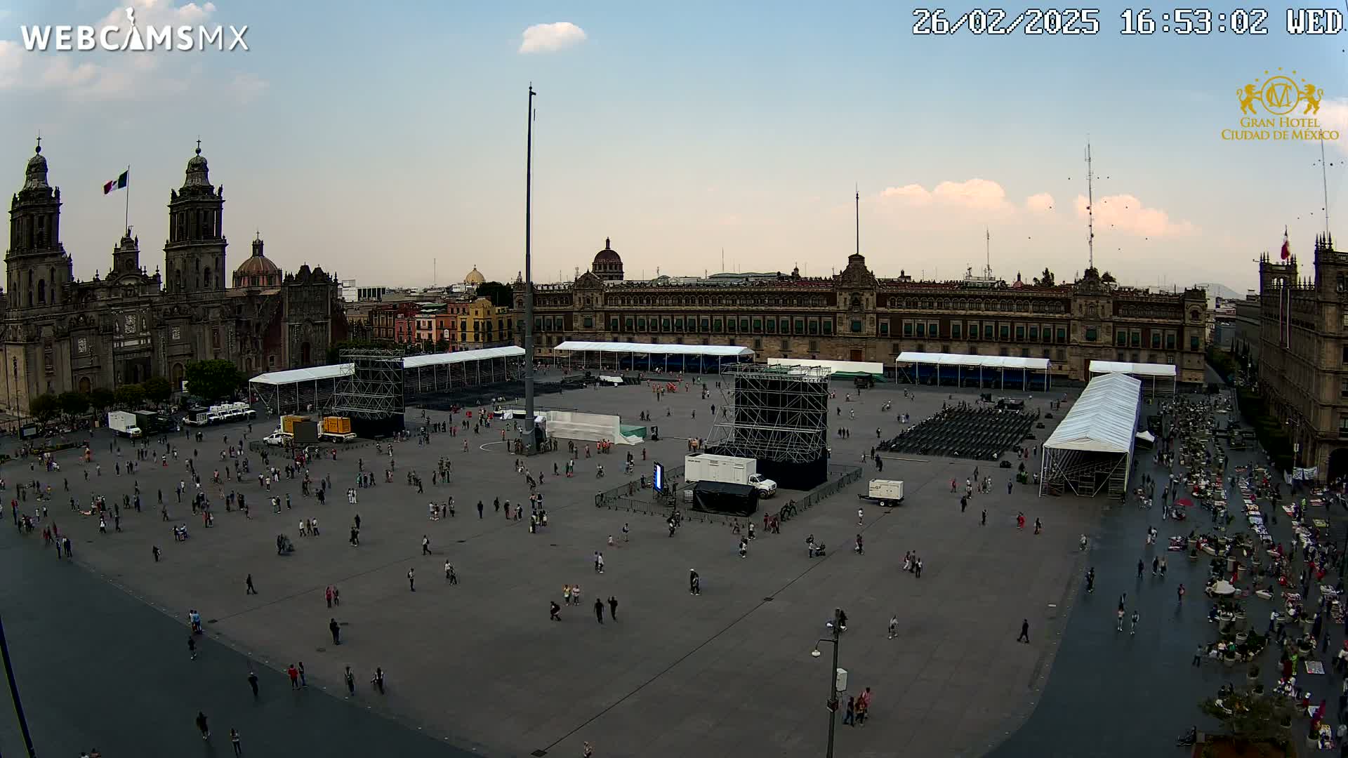 A large plaza with many people sparsely scattered across it, temporary structures, and large buildings including a cathedral in the background under a partly cloudy sky.
