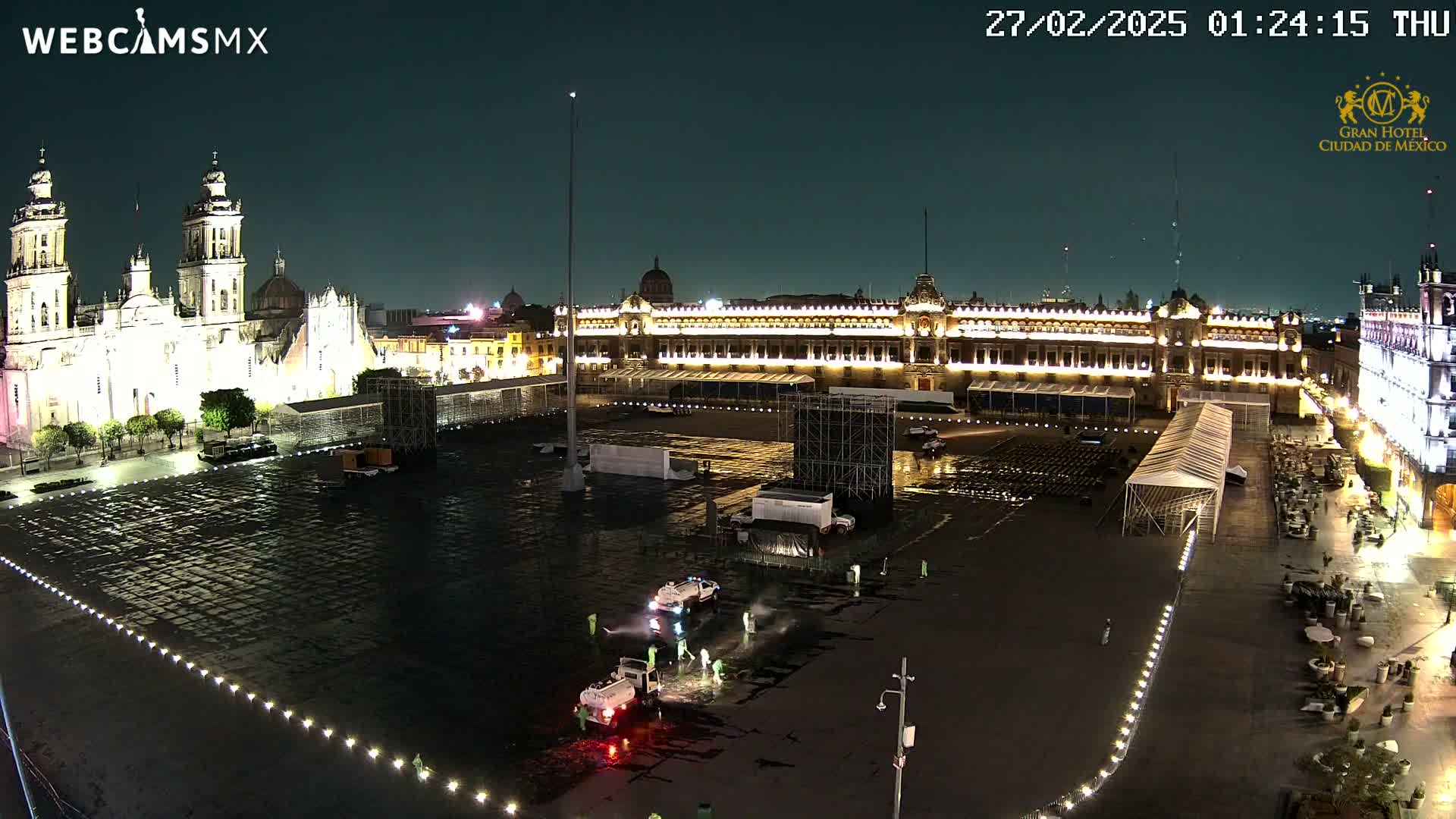 A large, mostly empty plaza at night is being cleaned by several workers and vehicles, with a brightly lit palace and cathedral in the background.