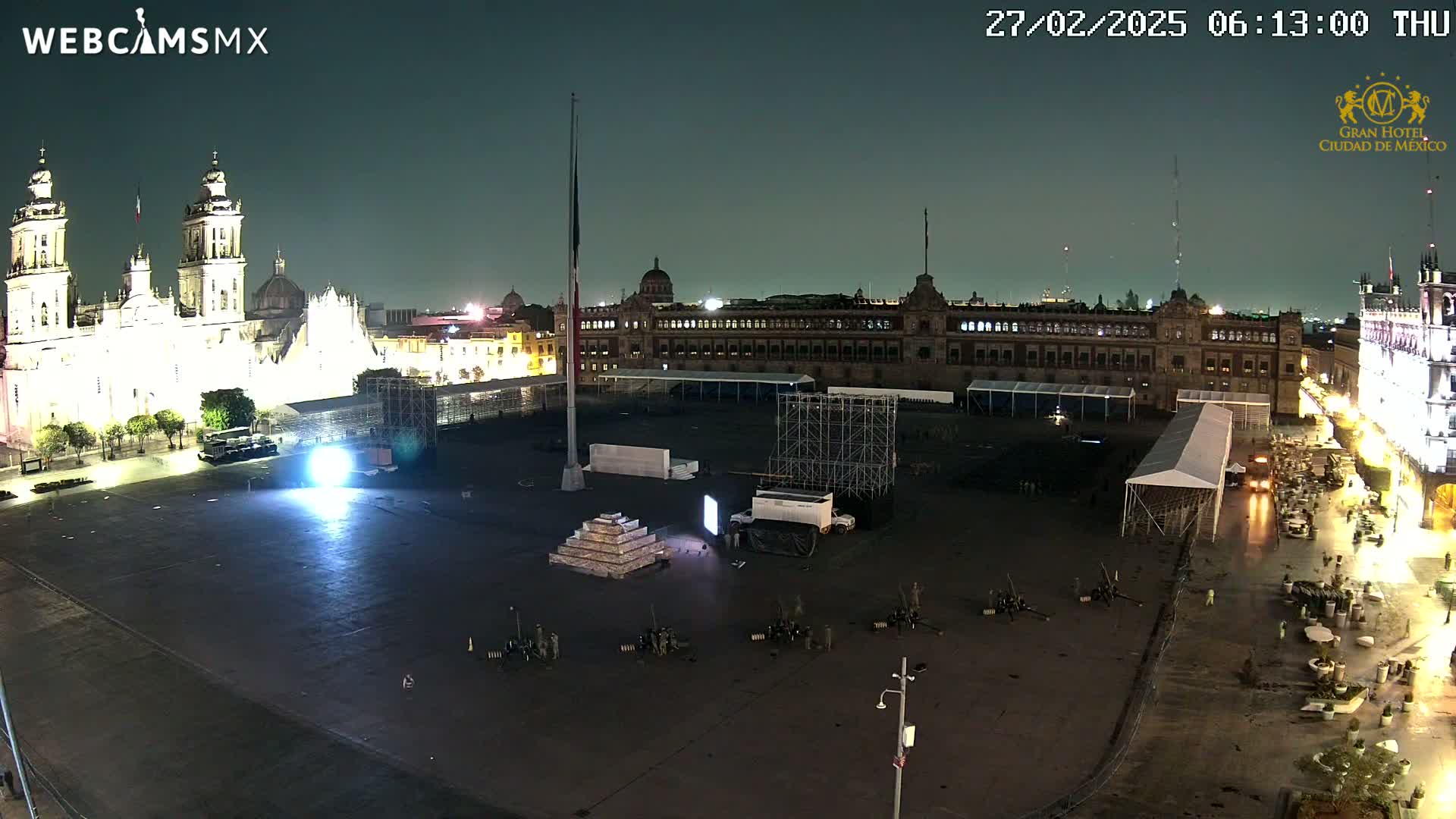 A nighttime, aerial view of a large plaza with a cathedral and government building illuminated, featuring a stage setup and several people, under a clear, dark sky.