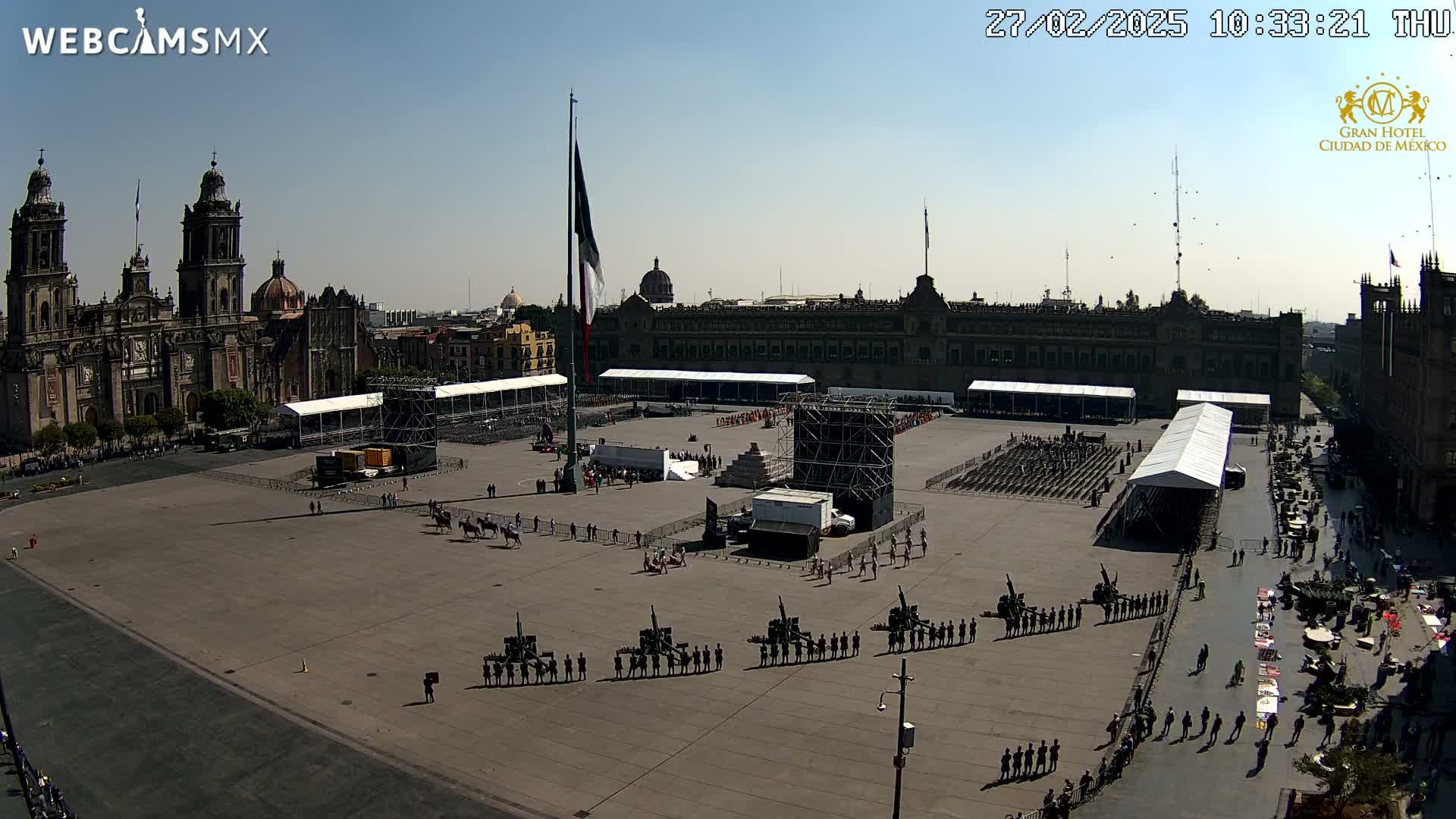 A large plaza, under a clear sunny sky, is being prepared for a large event, with military personnel in formation, several tents, and staging equipment visible.