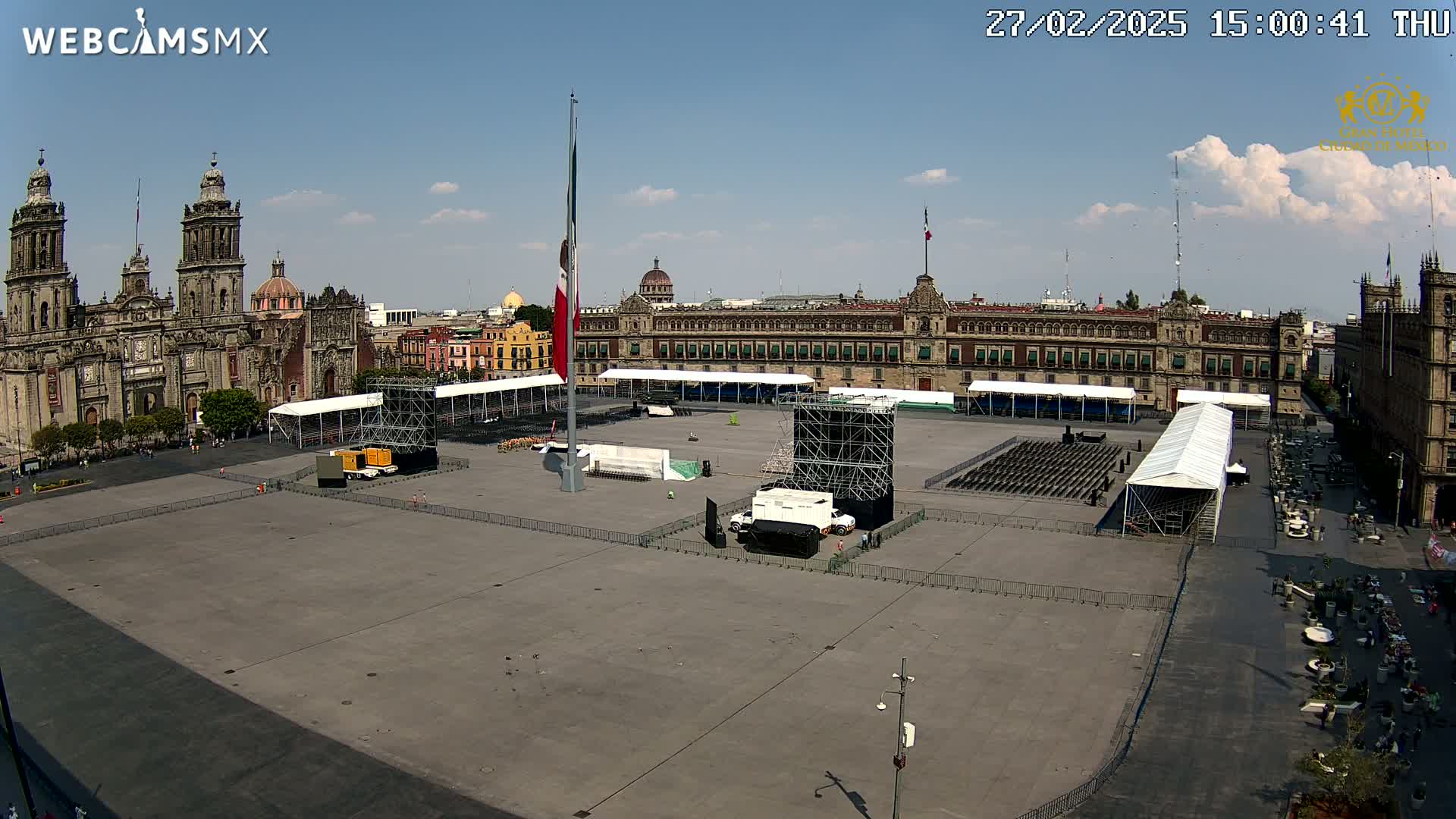 A large plaza, mostly empty except for staging equipment and seating, is situated in front of a large, ornate government building and a cathedral under a mostly sunny sky.