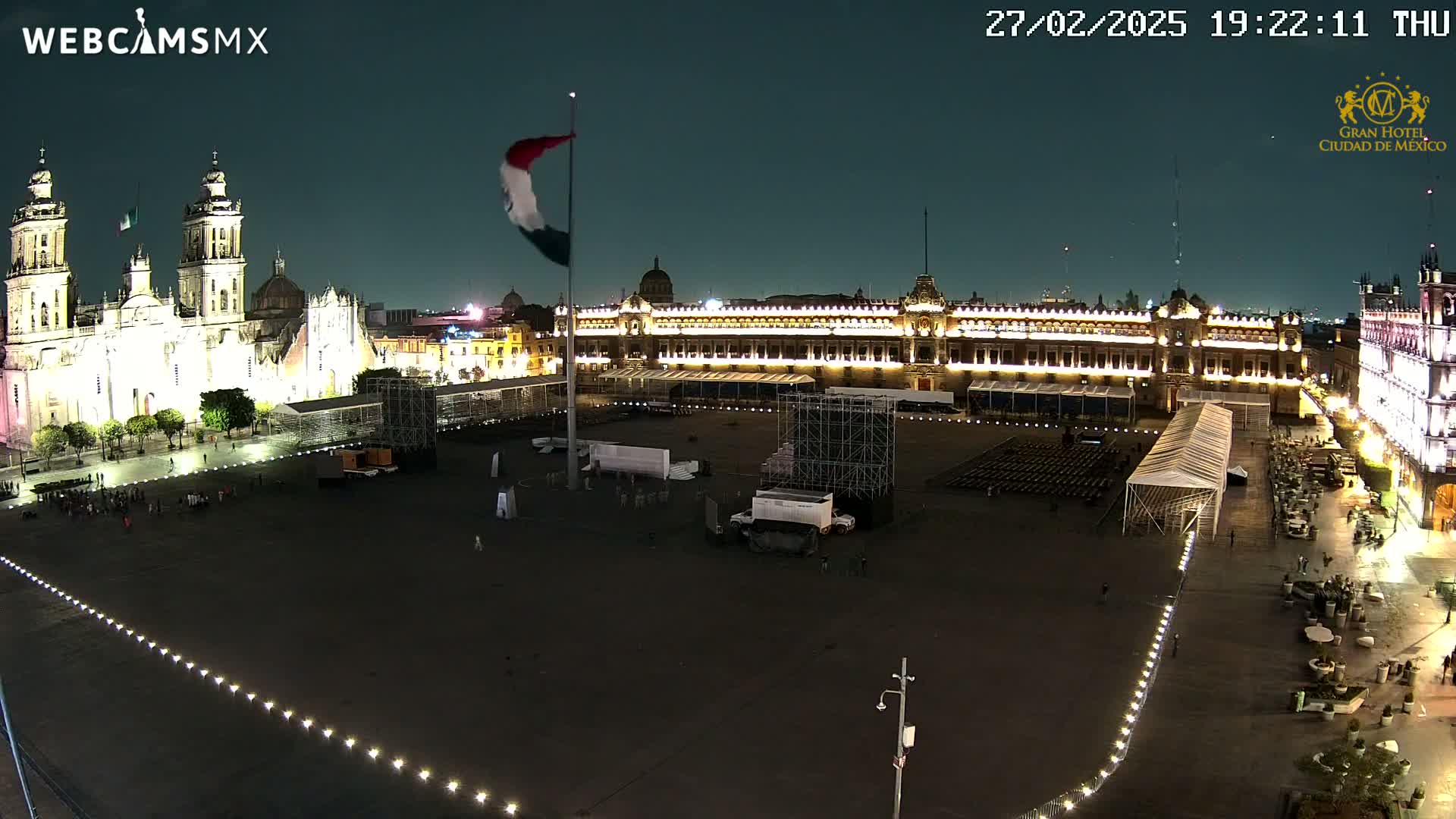 A nighttime, high-angle view of a large plaza with a cathedral and government building illuminated, surrounded by smaller buildings, and containing temporary event structures.