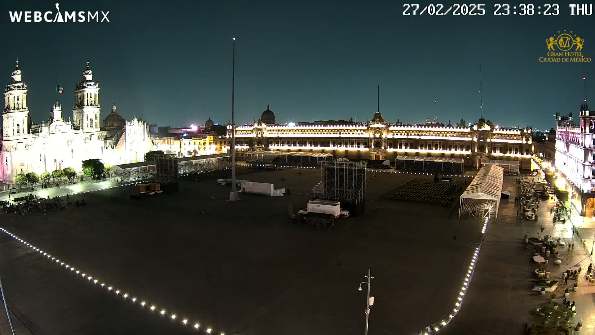 A nighttime, high-angle view of a large, mostly empty plaza with illuminated buildings, including a cathedral and a palace, surrounded by temporary structures and a few people.