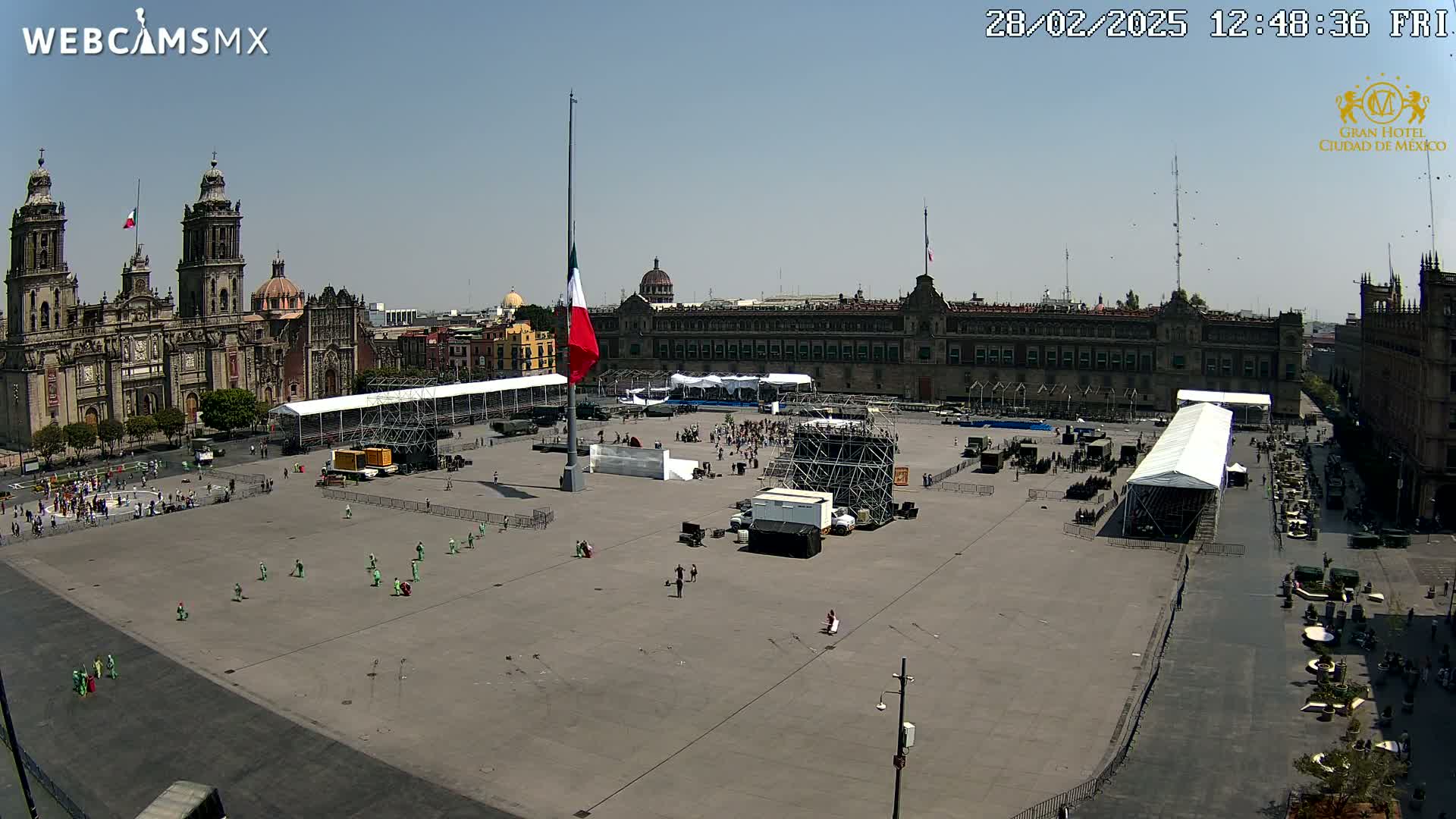 A large plaza is being prepared for an event, with temporary structures, fencing, and a few people present under a sunny sky.
