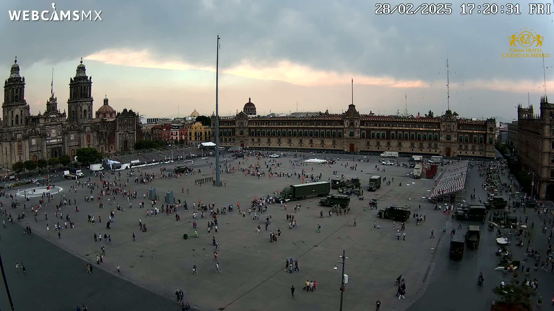A large plaza with many people and military vehicles under an overcast sky, surrounded by historical buildings including a cathedral and a palace.