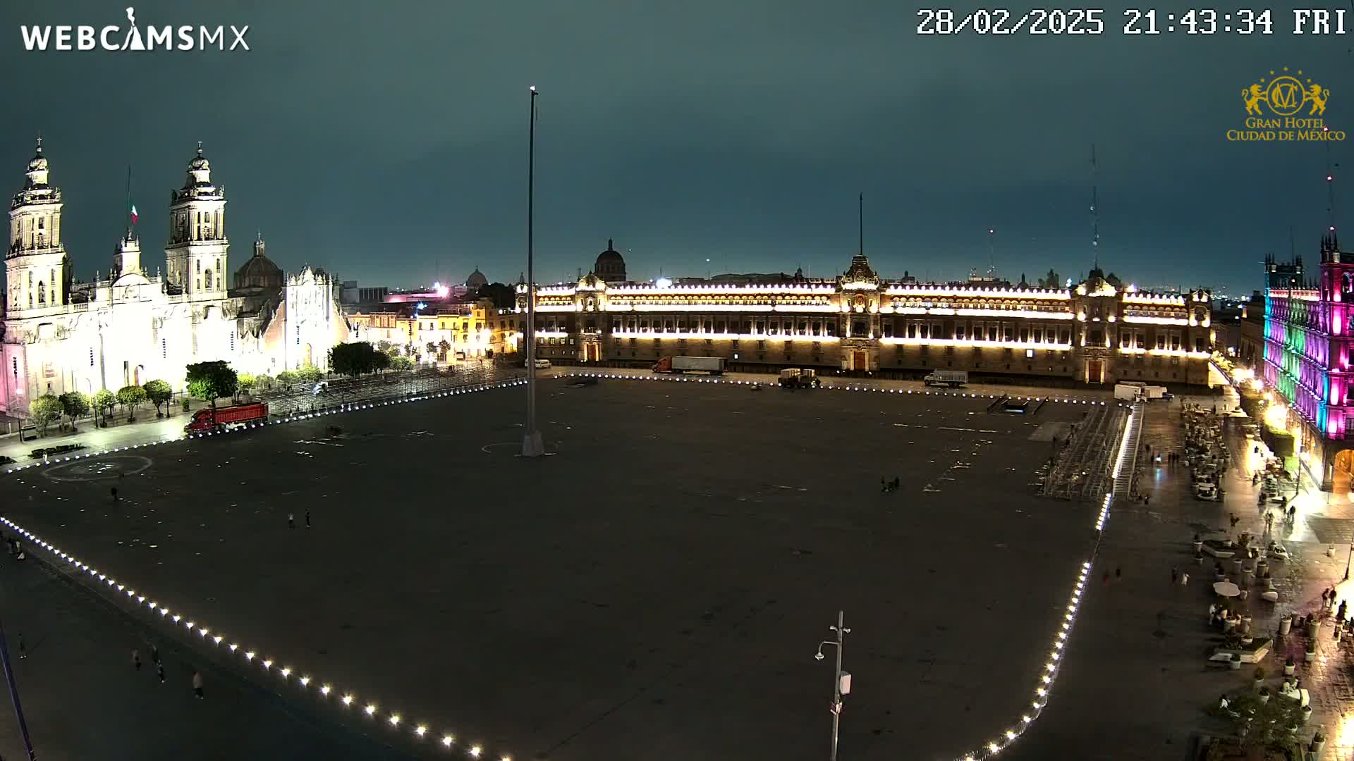 A large, mostly empty plaza at night is illuminated, with a brightly lit cathedral and government buildings visible in the background under a dark sky.