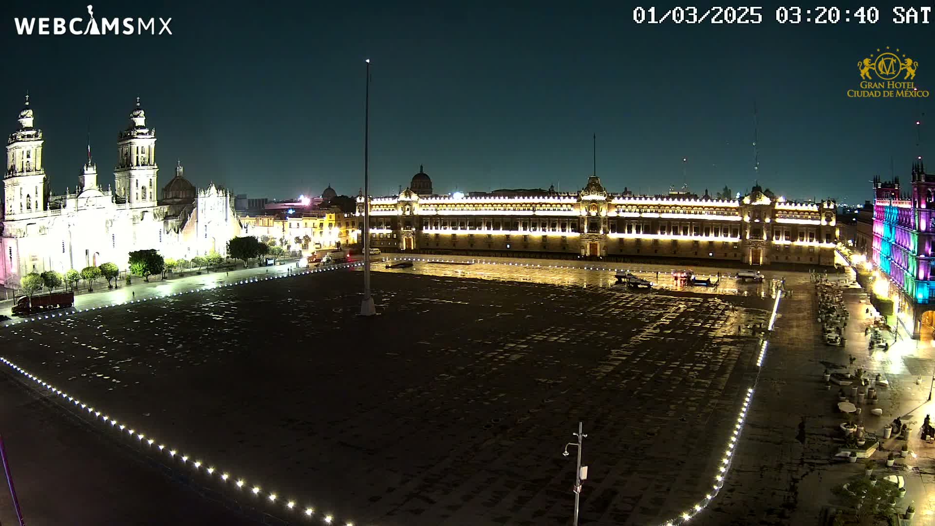 A large, empty plaza at night is illuminated, bordered by a brightly lit cathedral and palace, with a light rain falling.