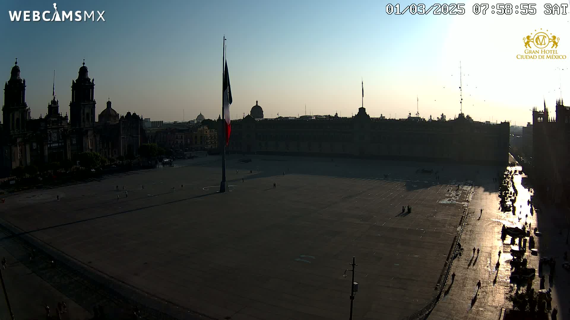A large plaza with a Mexican flag and cathedral in the background is mostly empty under a clear, sunrise sky.