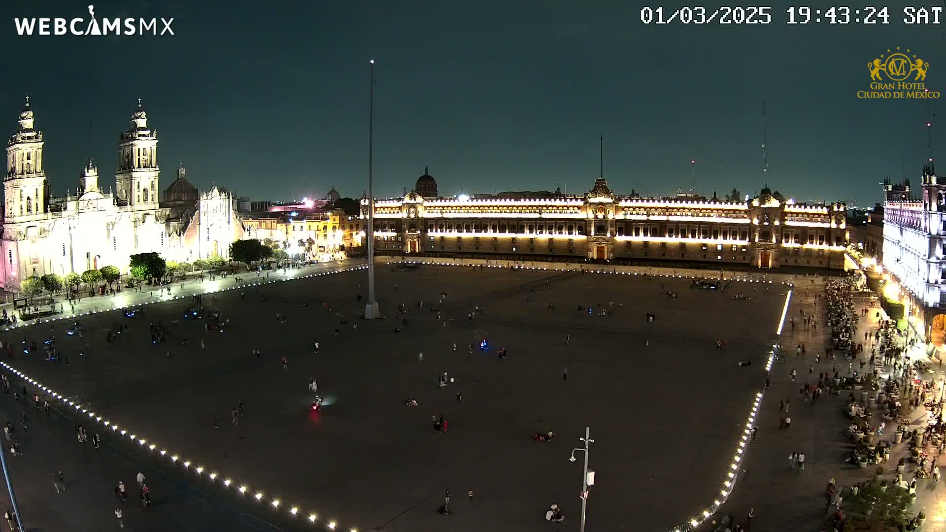 A large, dark plaza at night is illuminated by lights, with a cathedral and government building visible on either side, and many people scattered throughout the area.