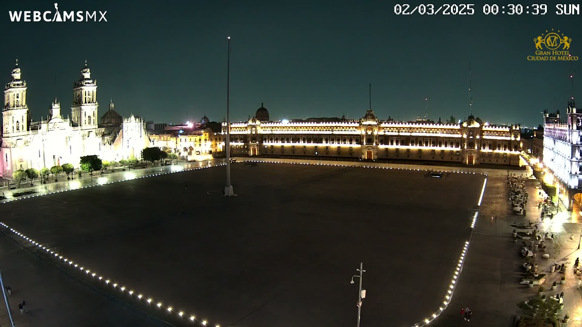 A large, mostly empty plaza at night is illuminated, with a cathedral and government buildings visible in the background under a dark sky.