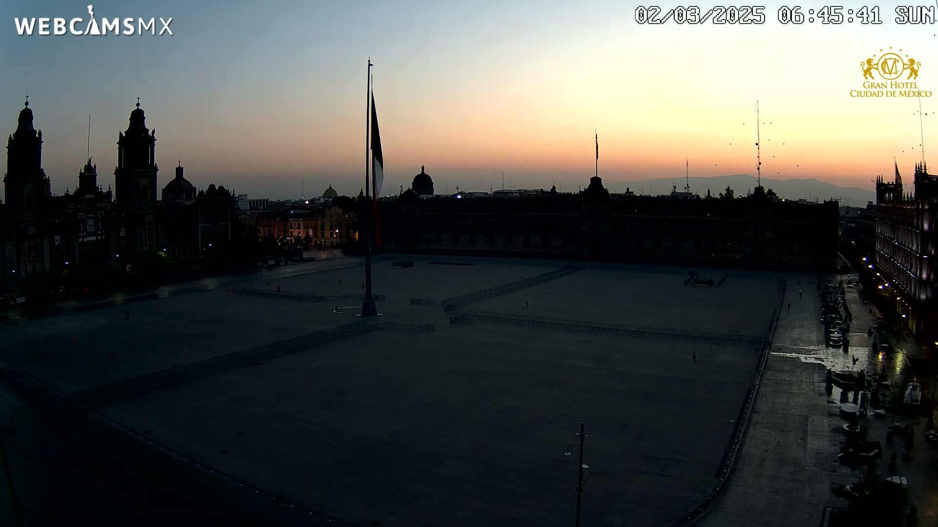 A large, dark plaza is mostly empty at sunrise, with a flagpole and surrounding colonial-era buildings silhouetted against the pale orange and blue sky.