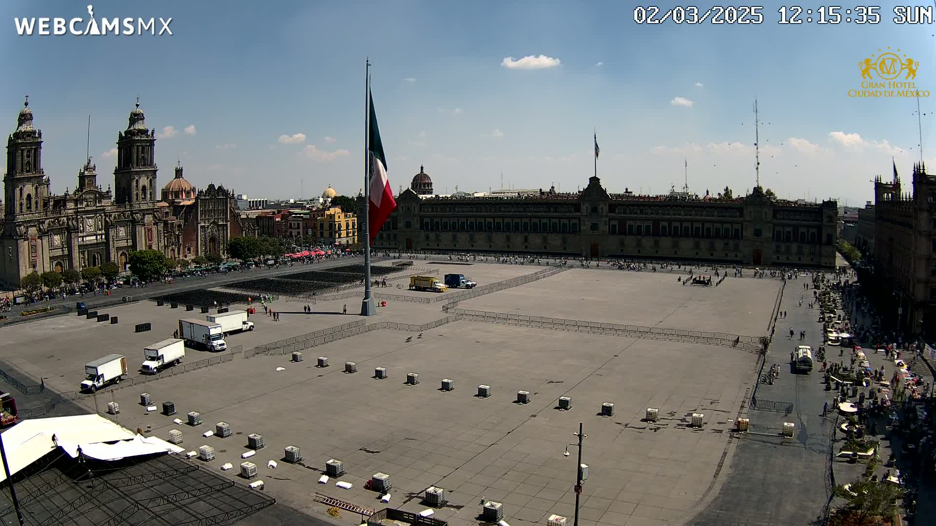 A large plaza with a Mexican flag, several trucks, and temporary fencing is seen under a sunny sky, with a large cathedral and government building in the background.