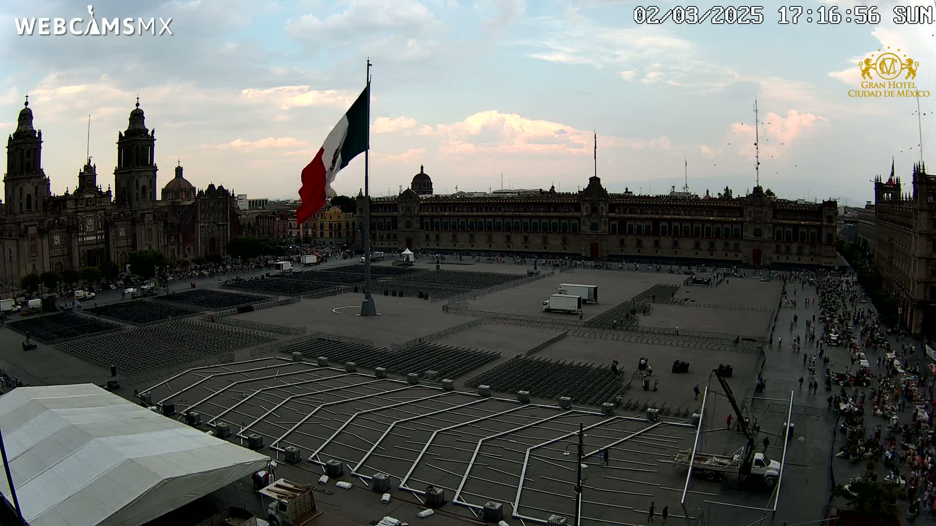 A large plaza with a Mexican flag, numerous empty chairs set up in rows, and several people scattered throughout, under a partly cloudy sky.