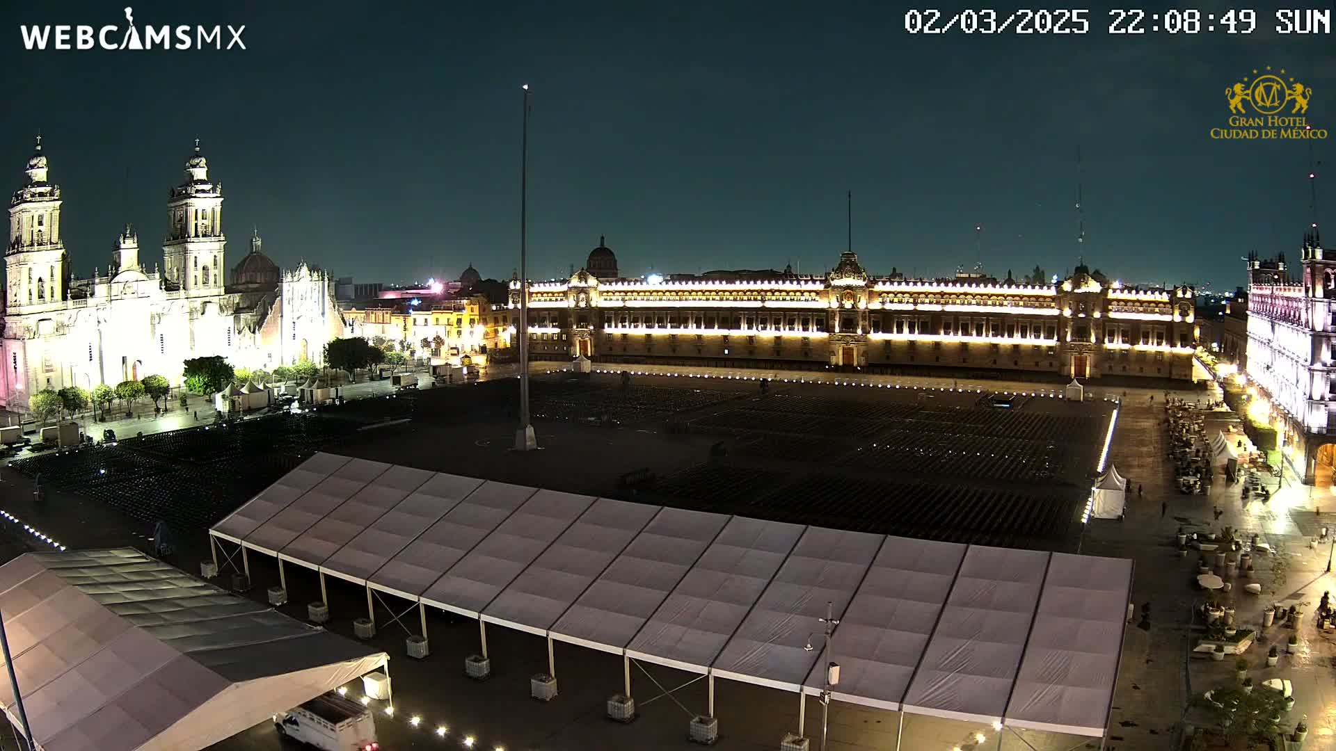 A nighttime view of a large, mostly empty plaza with a cathedral and government building illuminated, featuring several large tents in the foreground under a clear sky.