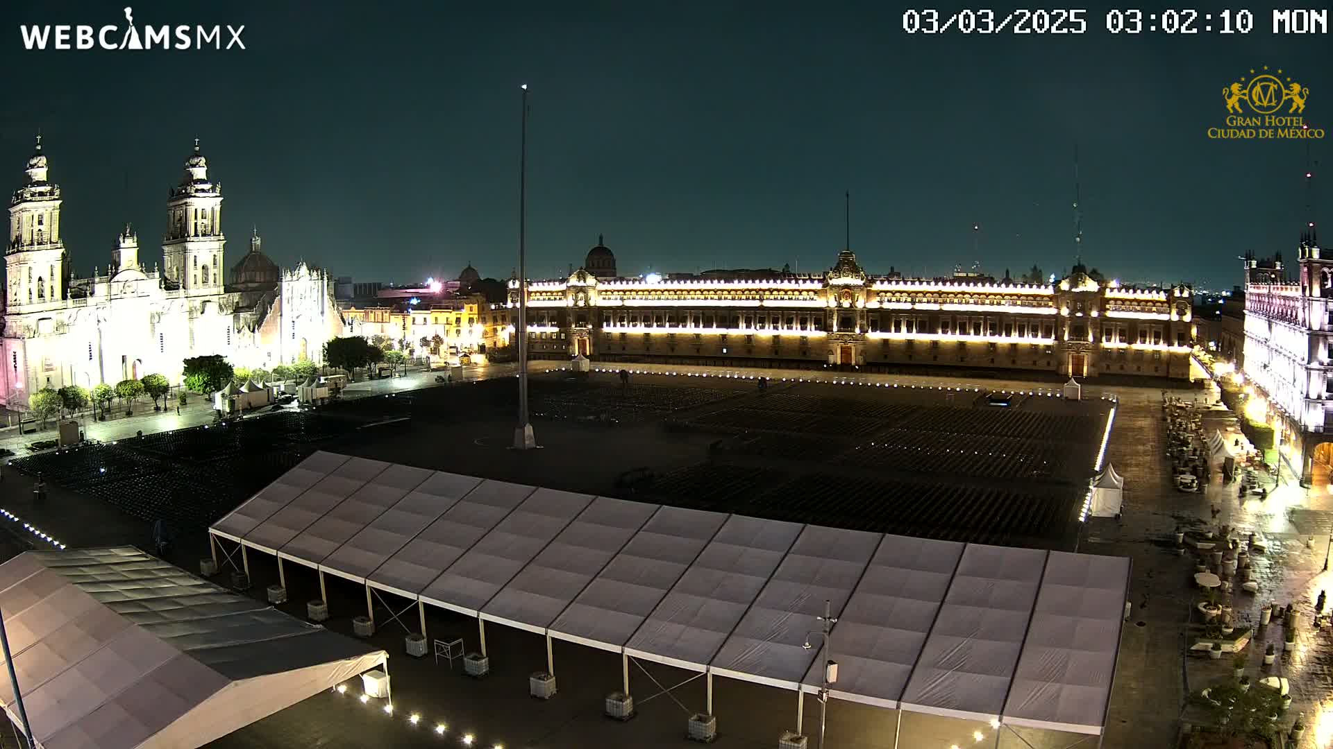 A large, mostly empty plaza at night, illuminated by lights, features a cathedral and government building, with several temporary tents set up.