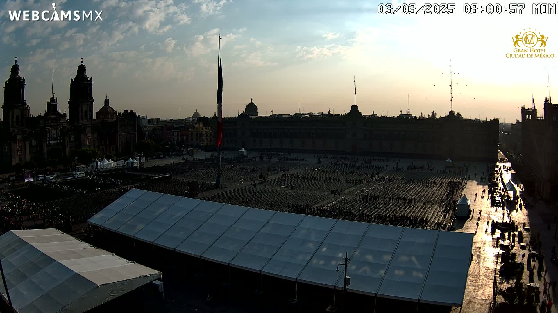 A large plaza with numerous people seated in rows under a partly cloudy sunset, surrounded by historical buildings and large tents.