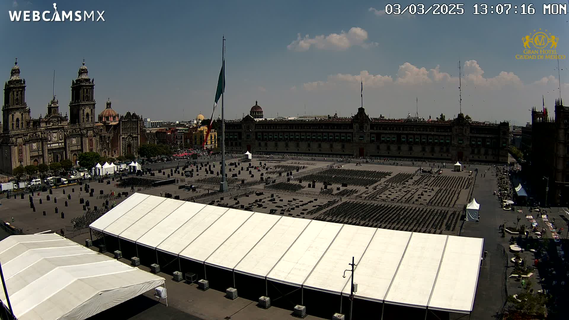 A large plaza, under a partly cloudy sky, is filled with numerous rows of chairs set up in front of a large building, with several white tents visible.