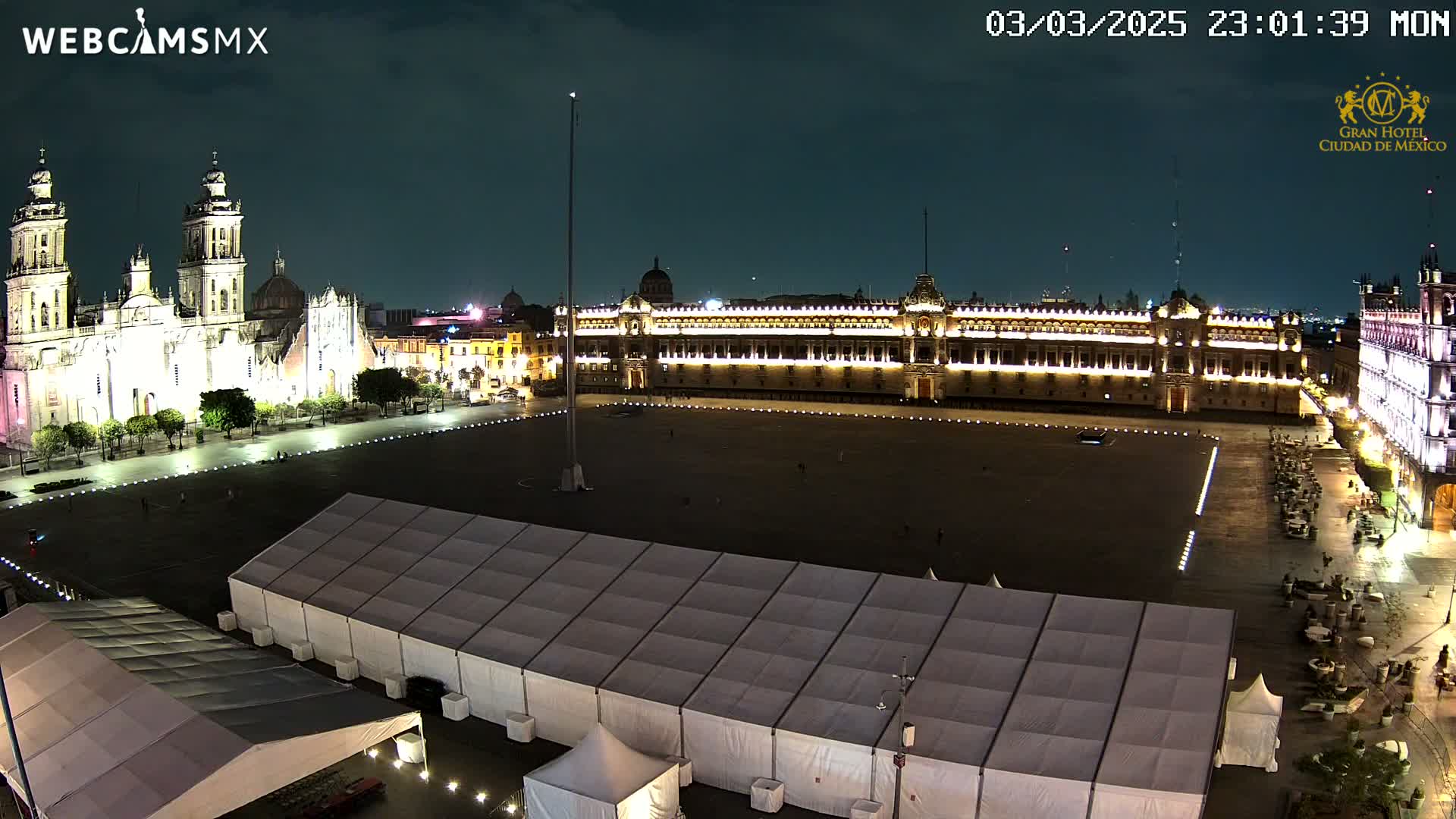 A large, mostly empty plaza at night is illuminated, showing a cathedral and government building, with several white tents set up in the foreground.