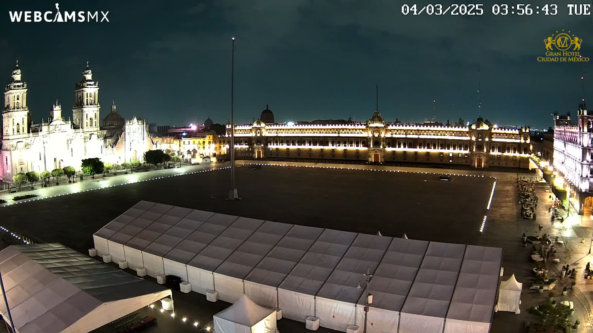 A large, mostly empty plaza at night, illuminated by lights, features a cathedral and government building in the background, with several white tents set up in the foreground under a partly cloudy sky.