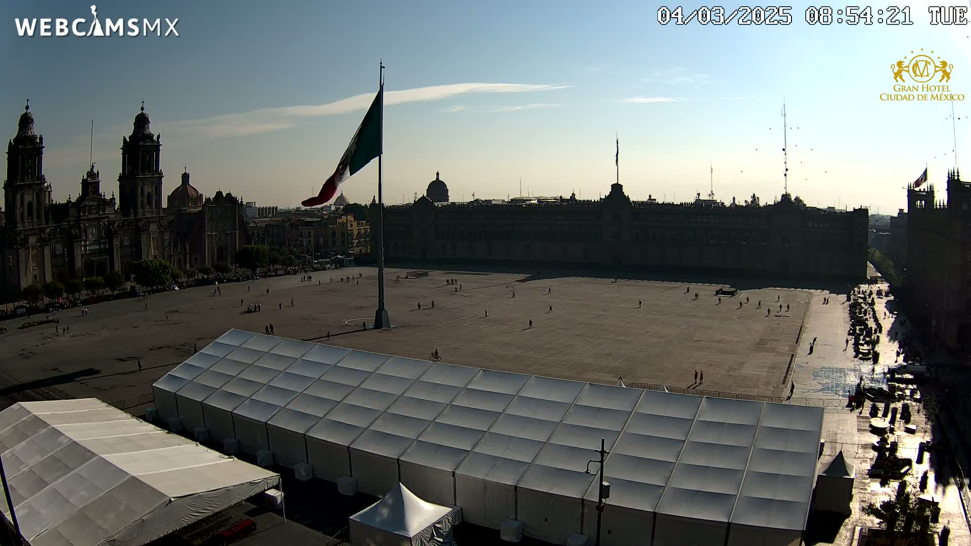 A large plaza with a Mexican flag, a cathedral, and a large building in the background under a mostly clear sky.