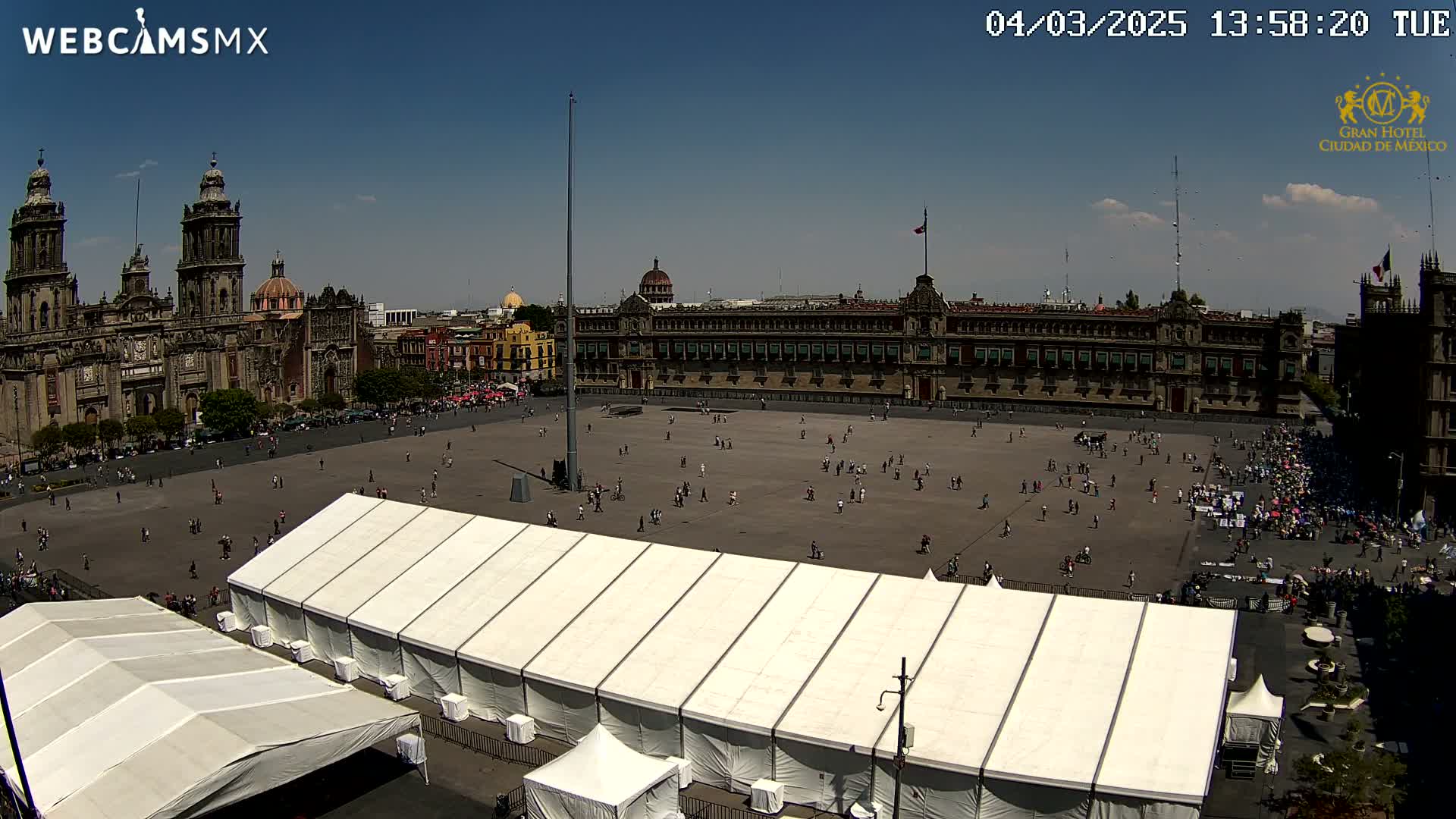 A large plaza with several people, a large white tent, and a large building in the background under sunny skies.