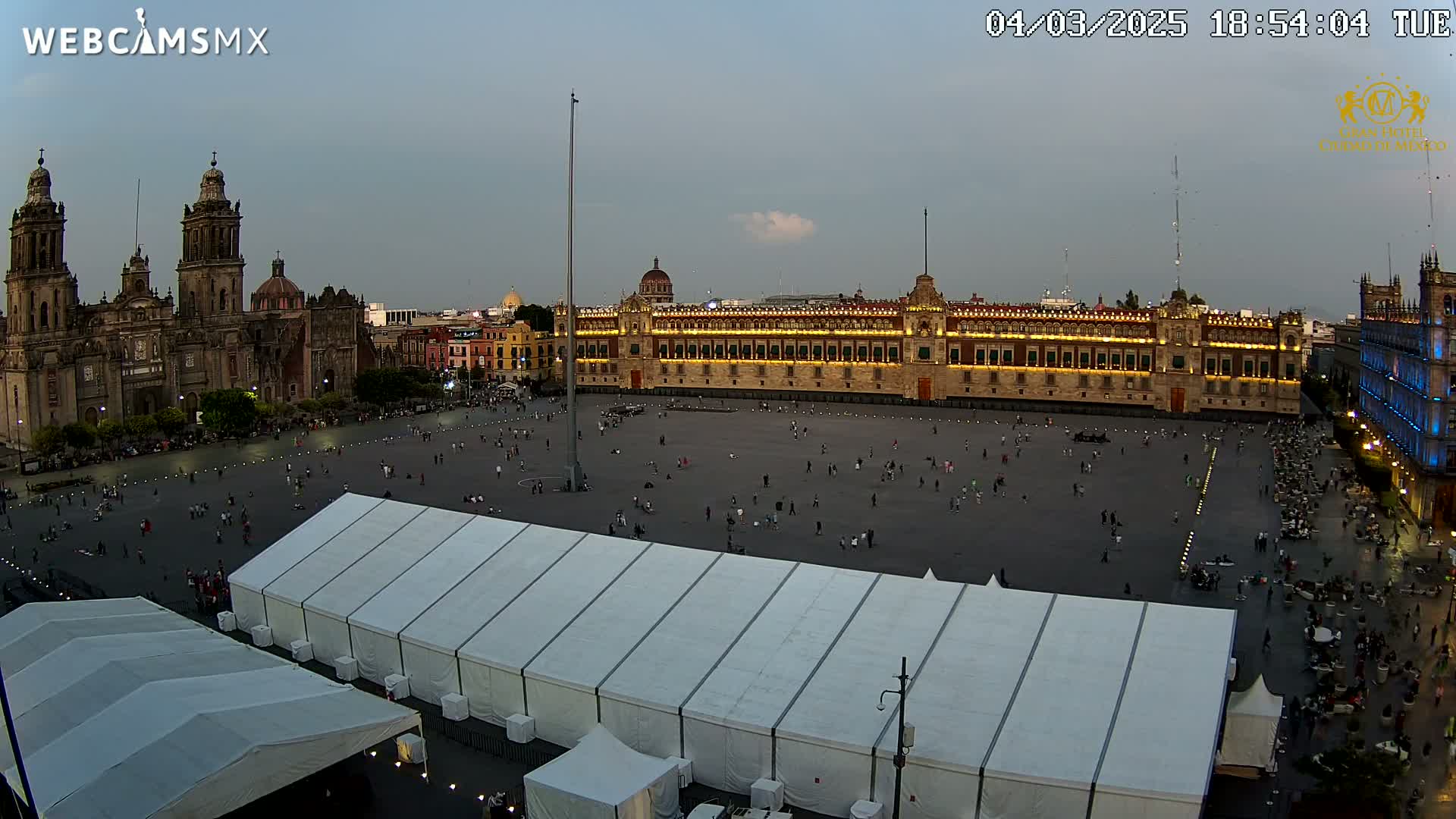 A large plaza at dusk, with a cathedral and government building visible, features several large white tents set up in the foreground.