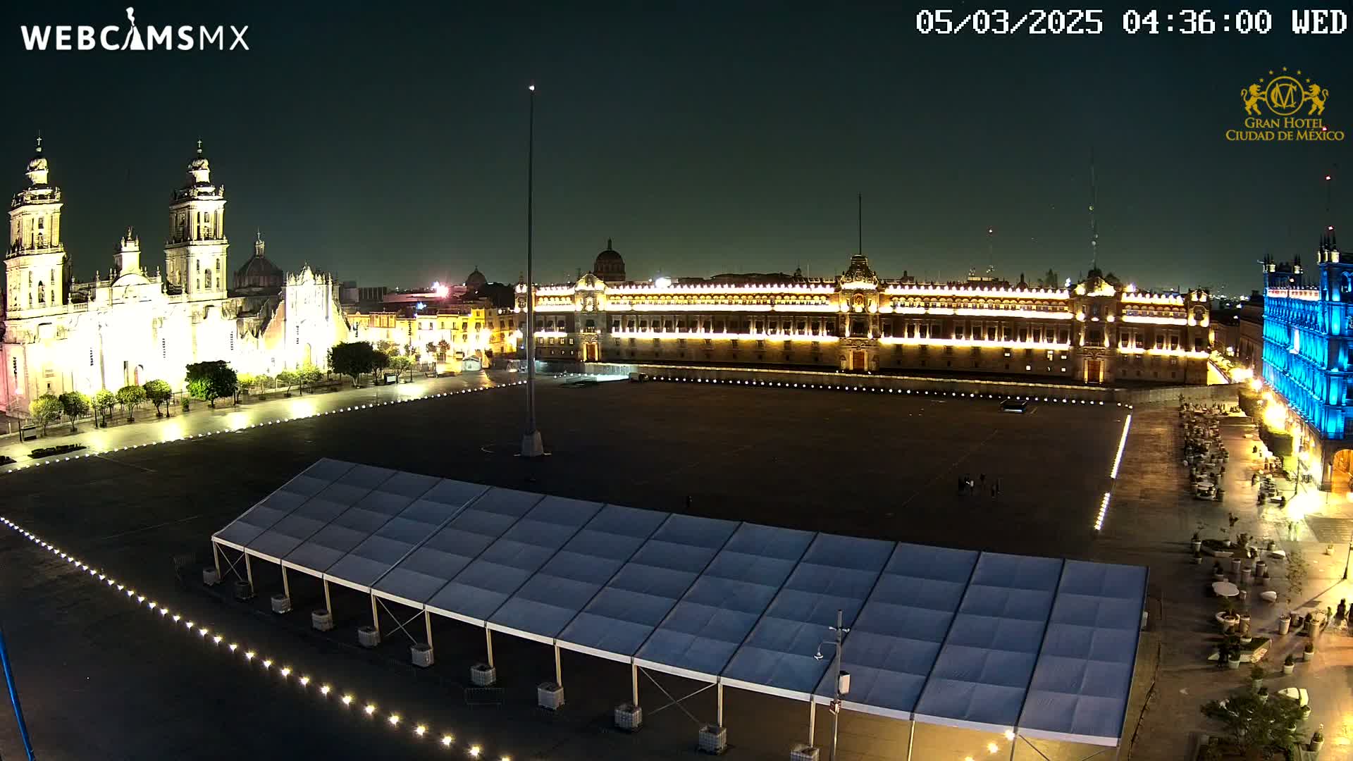 A large, empty plaza at night is illuminated, showing a cathedral and government building, with a long white tent in the foreground.