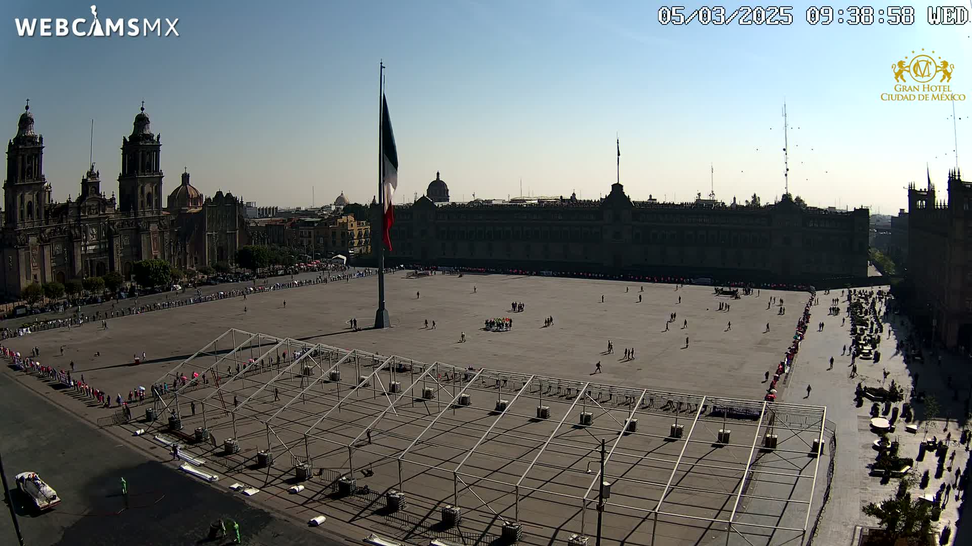A large plaza with a Mexican flag, temporary structures, and sparse crowds under a clear, sunny sky is overlooked by a cathedral and large buildings.