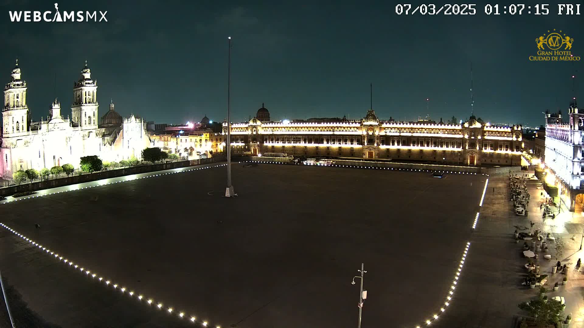 A large, dark plaza at night is illuminated by lights lining its edges, with a brightly lit cathedral and palace visible in the background under a clear night sky.