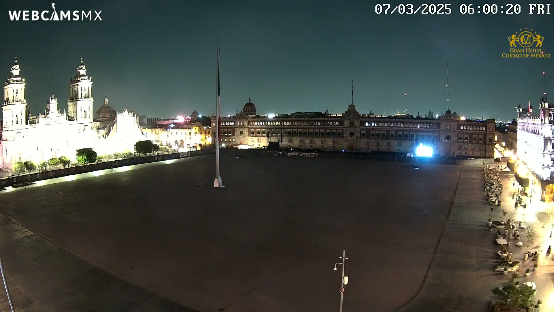 A large, mostly empty plaza at night is illuminated, with a brightly lit cathedral and government building visible in the background under a dark sky.