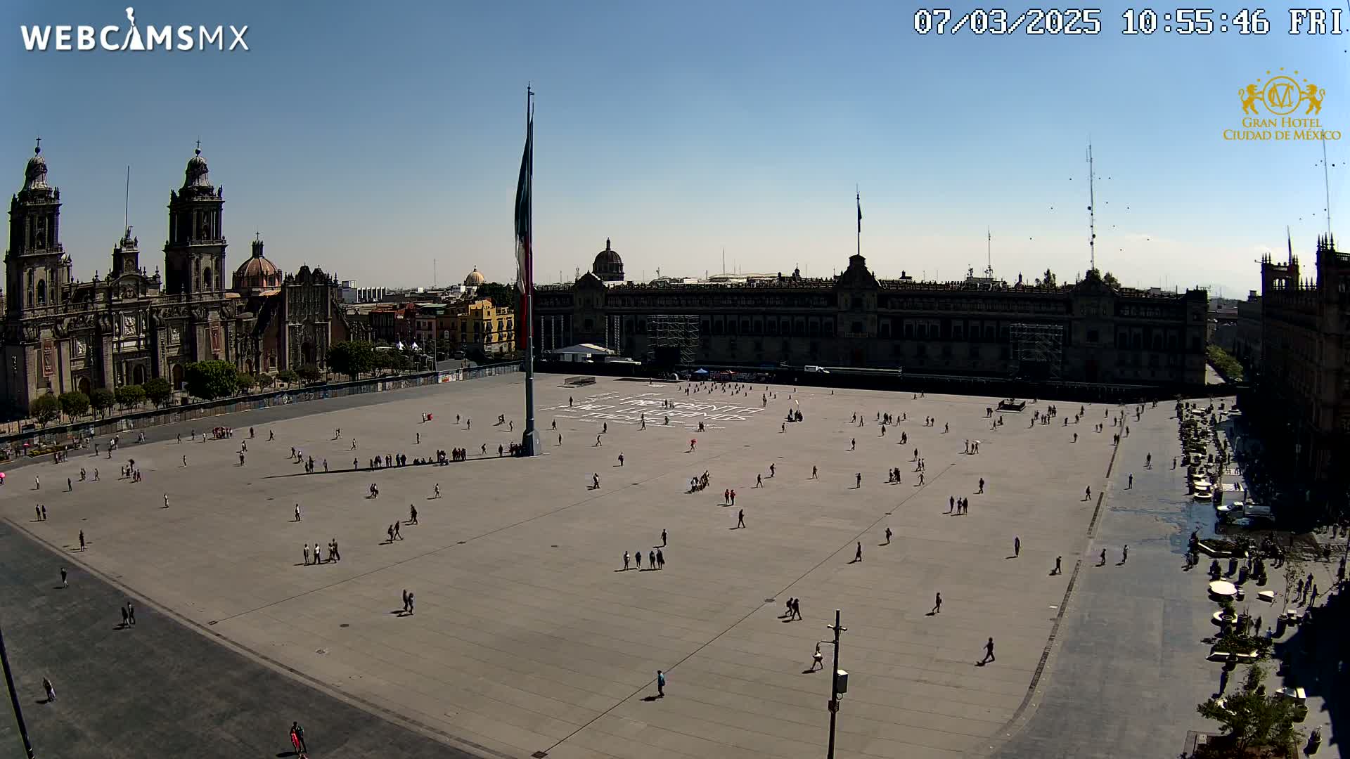 A large plaza with scattered people is seen under a clear, sunny sky, with a cathedral and government buildings in the background.