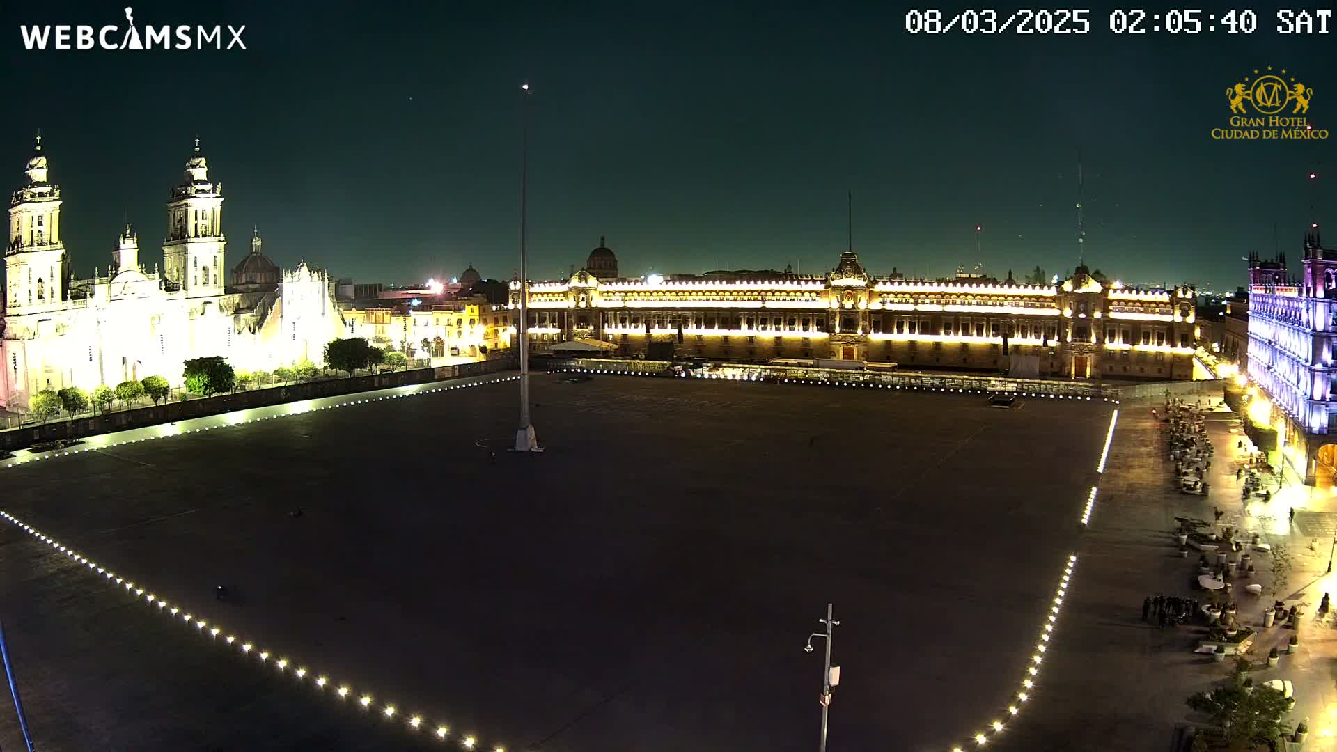 A large, mostly empty plaza at night, illuminated by lights, is flanked by a brightly lit cathedral and government building.