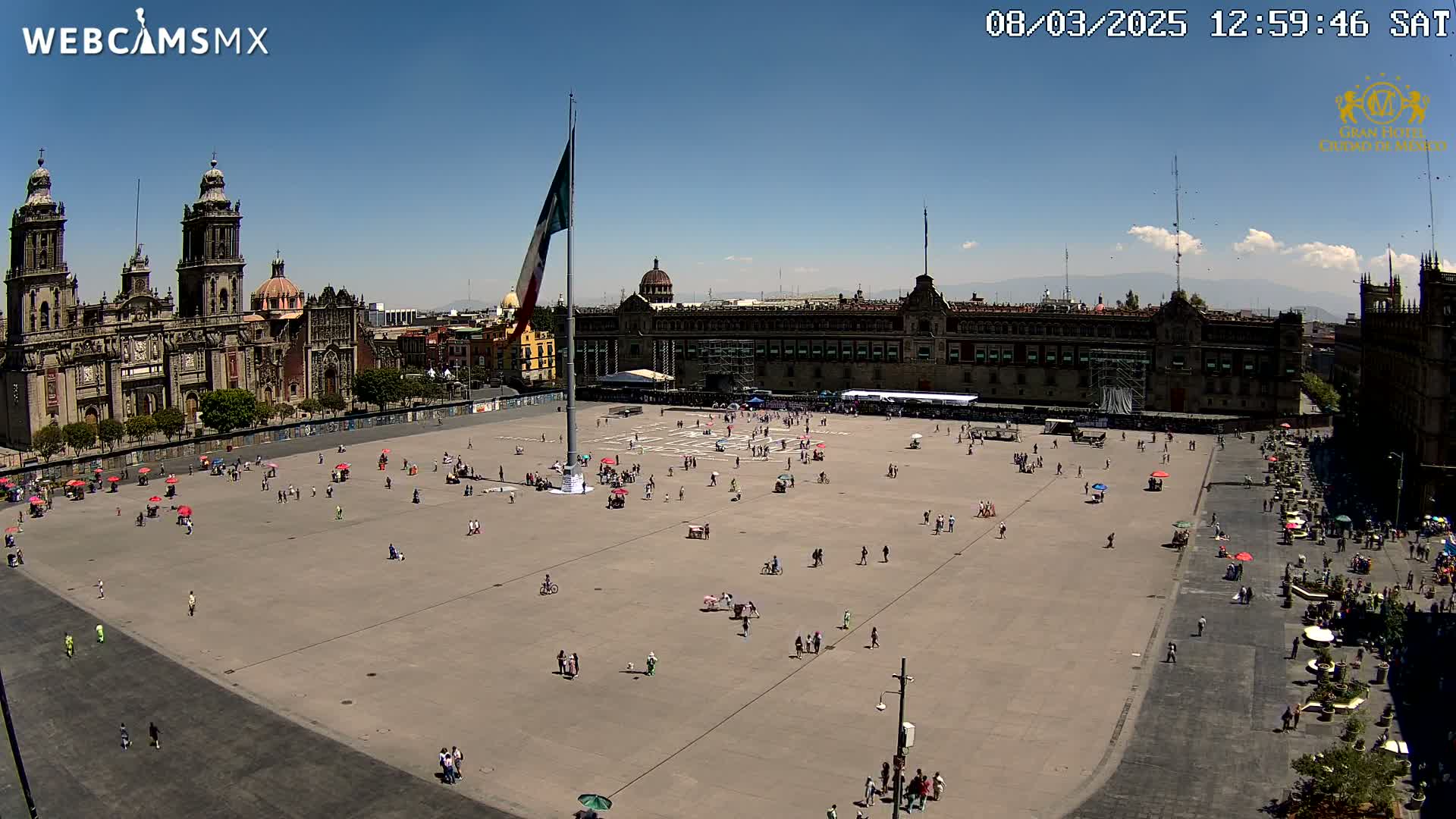 A large plaza with a few people scattered throughout, a large flagpole in the center, and a grand cathedral and palace visible in the background under a mostly sunny sky.
