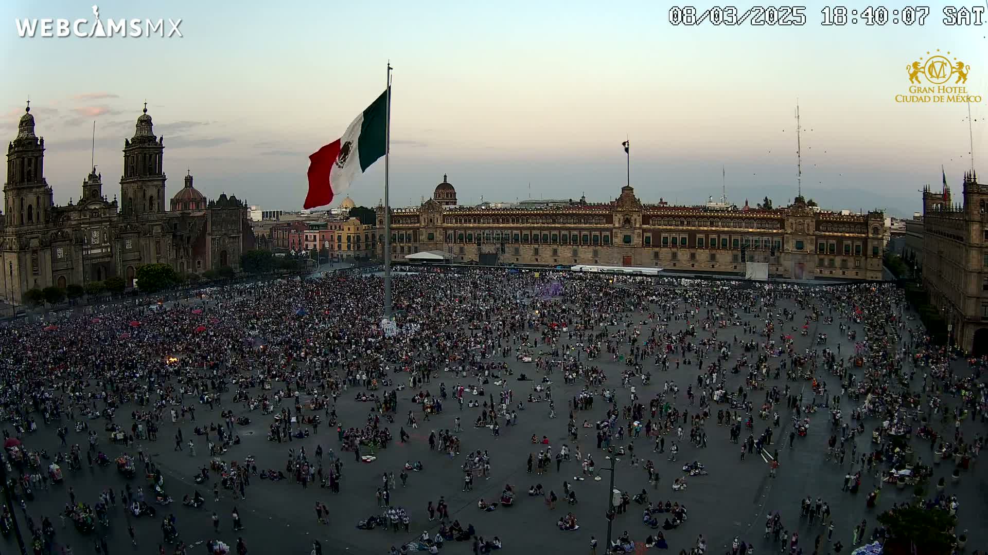 A large crowd gathers in a plaza at dusk, in front of a cathedral and a large government building, under a mostly clear sky.