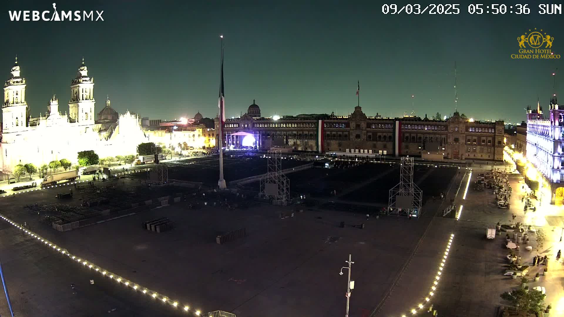 A large, mostly empty plaza at night is lit by artificial lights, with a cathedral and government buildings visible in the background.