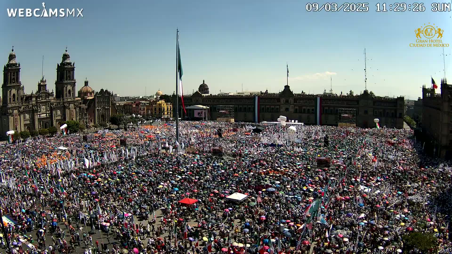 A large crowd gathers in a plaza in front of a cathedral and government buildings under a clear, sunny sky.