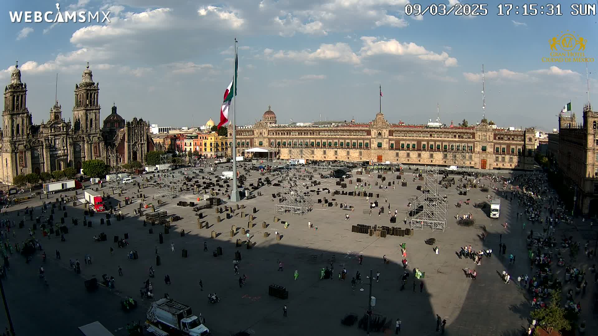 A large plaza, partially filled with numerous dark rectangular objects, is situated between a cathedral and a large government building under a partly cloudy sky.