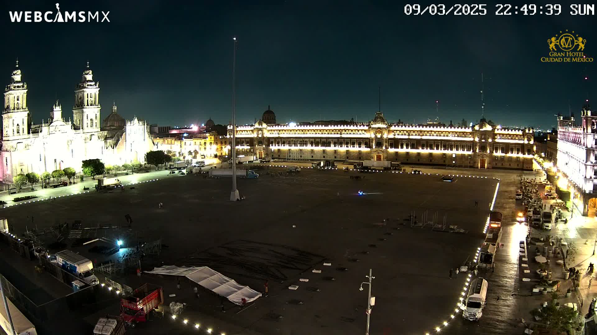 A large, mostly empty plaza at night is illuminated, showing a cathedral and government buildings, with several trucks and staging equipment present.
