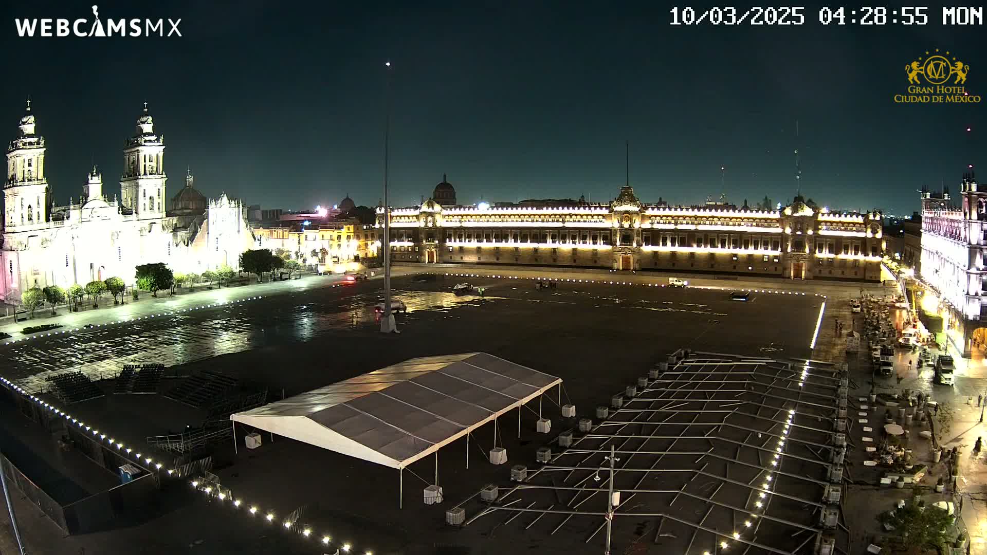 A large, empty plaza at night, illuminated by lights, shows a cathedral and government building, with a temporary tent and setup in the foreground under a clear, dark sky.