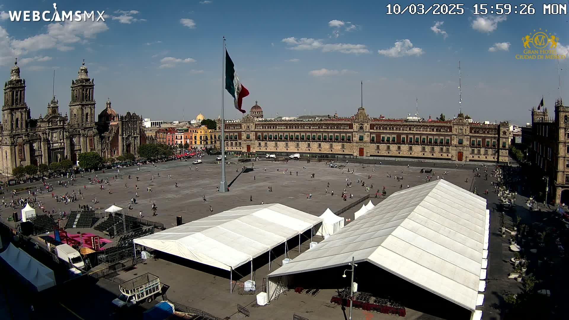 A large plaza with several white tents, a cathedral, and a government building under a mostly sunny sky.