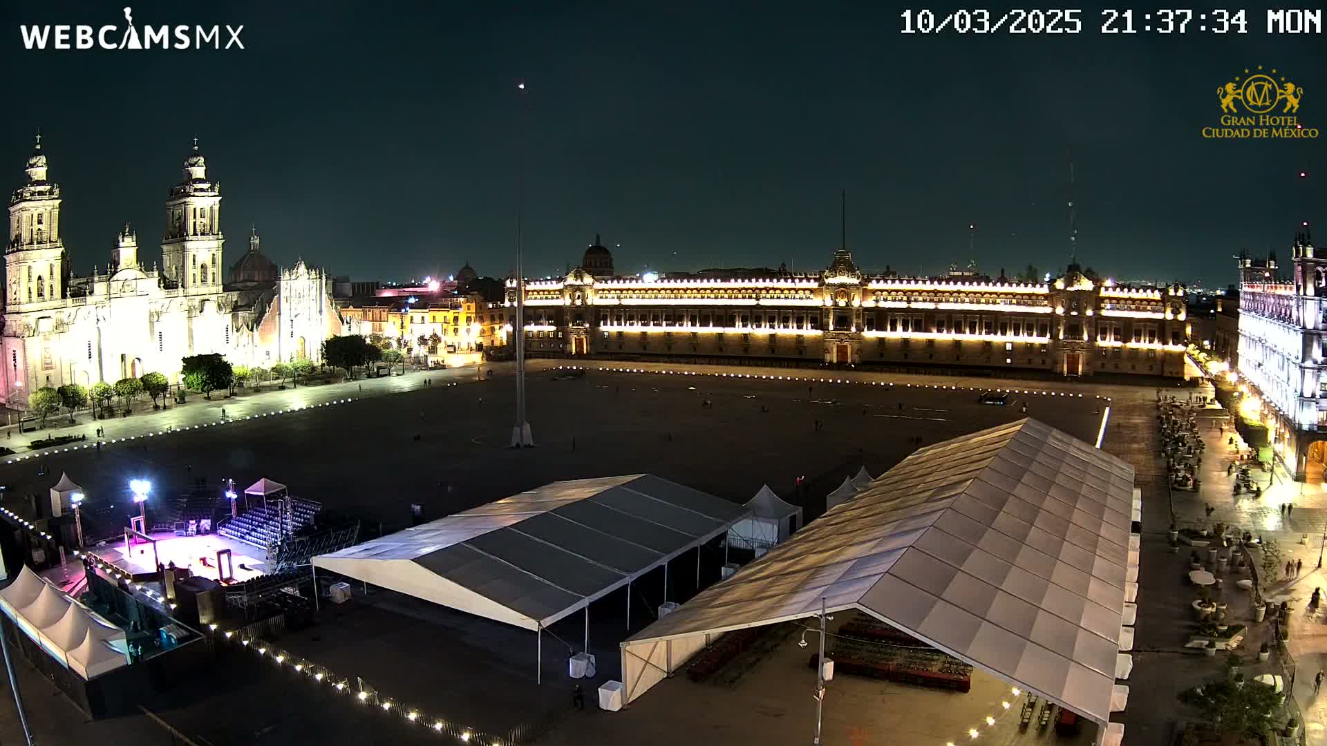 A brightly lit plaza at night features a large cathedral and government building, with several temporary tents set up in the foreground under a clear, dark sky.