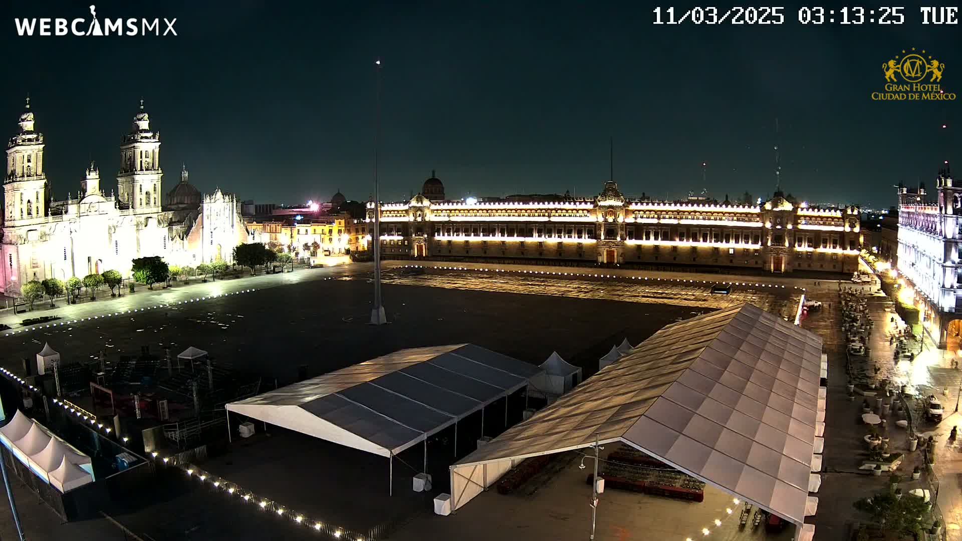 A nighttime, aerial view of a large plaza with a cathedral and government building illuminated, several tents set up in the plaza, and a few people scattered around.