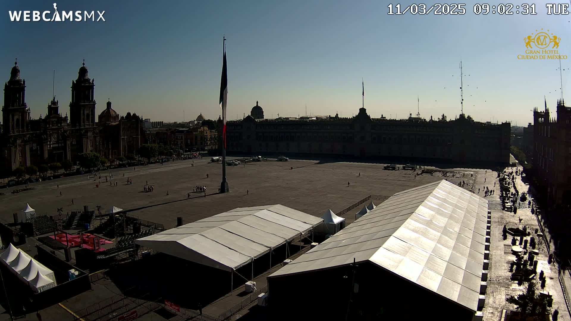 A large plaza, under a clear sunny sky, features several large white tents and is surrounded by historic buildings, including a cathedral and a large government building.