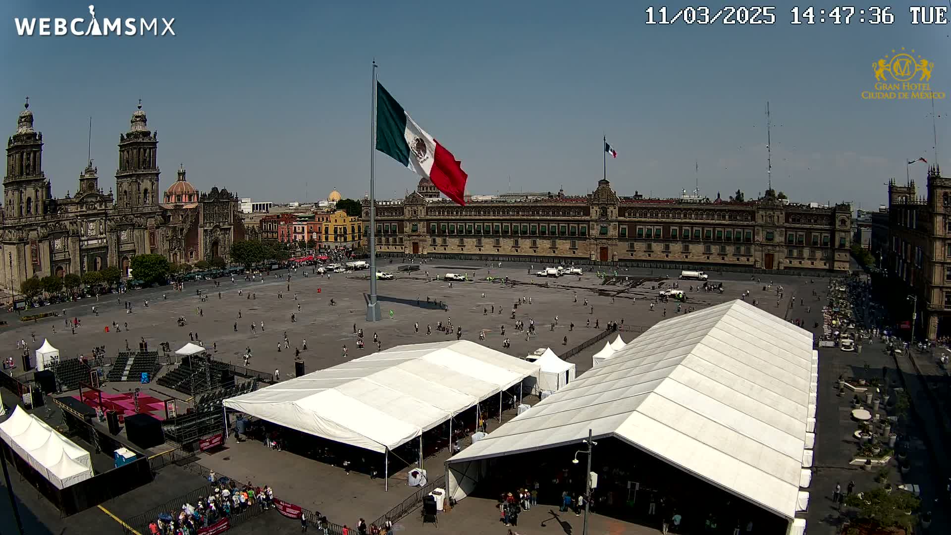 A large plaza with several white tents, a tall flagpole flying the Mexican flag, and a large cathedral in the background under a clear, sunny sky.